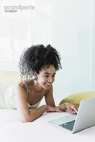 Glückliche schöne Frau  die auf dem Bett liegt und zu Hause einen Laptop benutzt