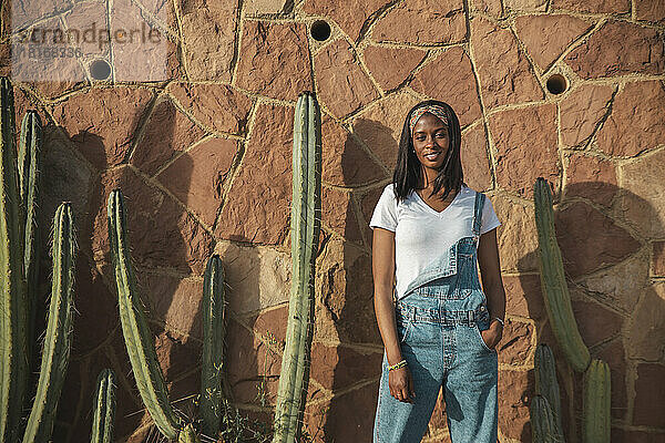 Selbstbewusste junge Frau steht vor einer Wand mit Kaktuspflanze
