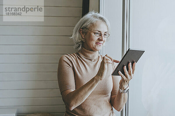 Lächelnde Frau mit Tablet-PC  die zu Hause am Fenster steht