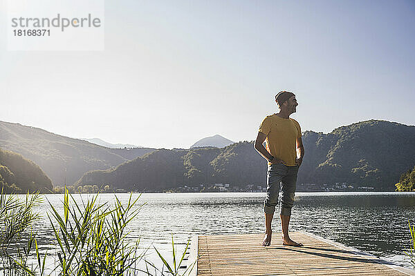 Lächelnder Mann mit Strickmütze und Händen in den Taschen auf dem Steg über dem See