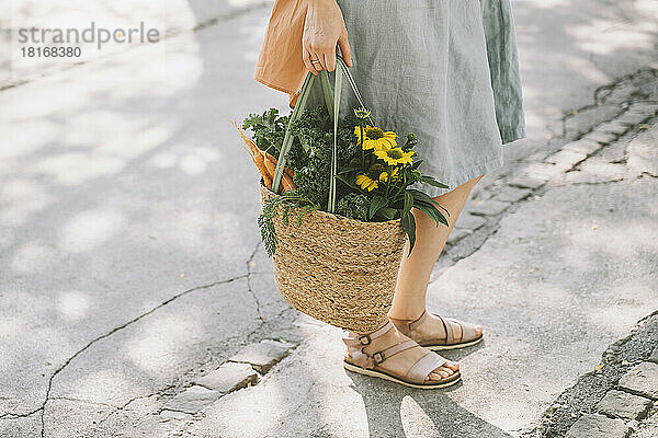 Frau steht auf der Straße und hält eine Tüte mit Gemüse und Blumen in der Hand