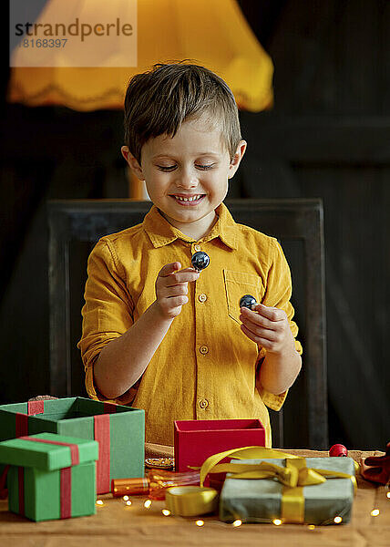 Lächelnder Junge mit Weihnachtskugeln und Geschenken am Tisch