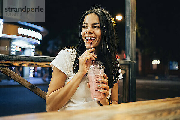 Fröhliche Frau mit Smoothie sitzt nachts im Straßencafé