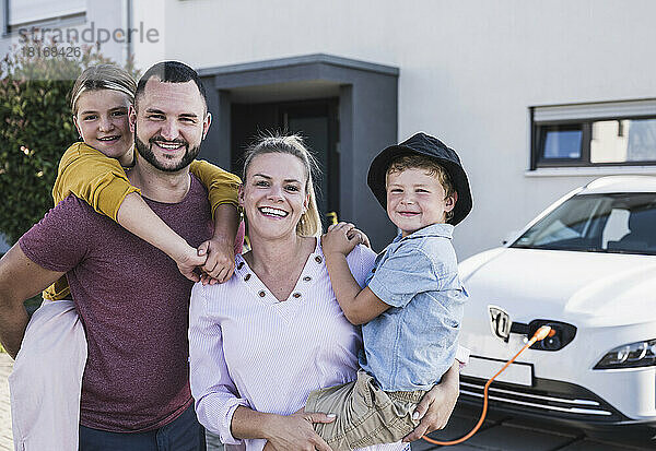 Glückliche Familie steht vor dem Einfamilienhaus und dem Elektroauto