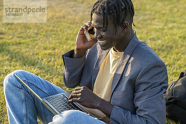 Glücklicher junger Geschäftsmann  der am Handy sitzt und mit einem Laptop spricht
