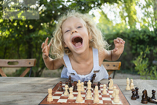 Aufgeregtes Mädchen mit Schachbrett auf Tisch im Garten