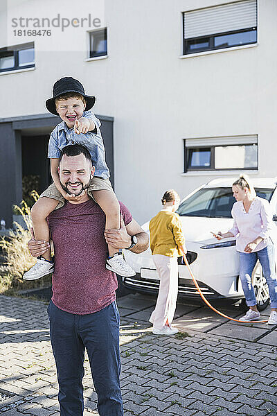 Glücklicher Vater trägt Sohn auf Schultern  während Mutter und Tochter Elektroauto aufladen