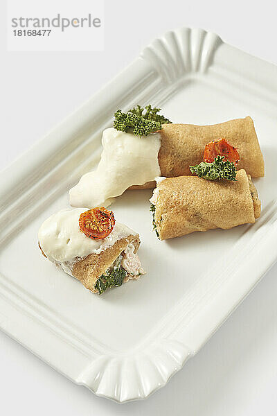 Stuffed shrimp crepes garnished with tomato and cream arranged in tray against white background