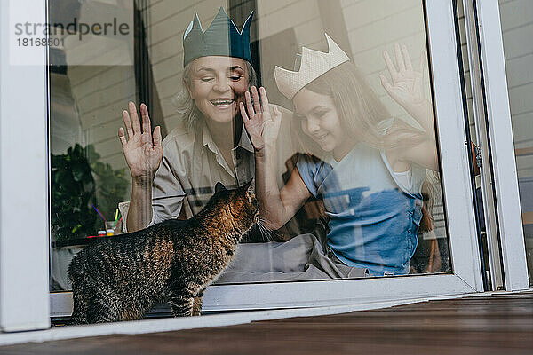 Glückliche Großmutter und Enkelin mit Krone und Blick auf die Katze durch das Glasfenster