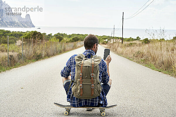 Mann mit Rucksack und Smartphone sitzt auf Skateboard