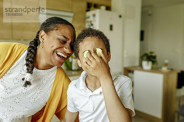 Glückliche Mutter umarmt ihren Sohn und bedeckt zu Hause die Augen mit Gurken