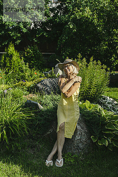 Frau umarmt sich selbst und lehnt an einem Felsen neben Pflanzen im Garten