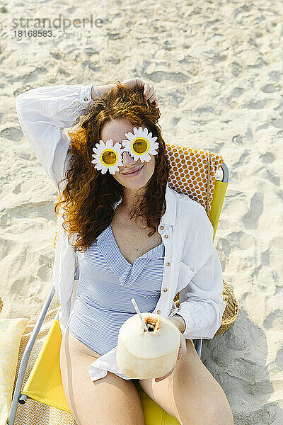 Lächelnde Frau mit Blumensonnenbrille sitzt mit Kokosnuss am Strand