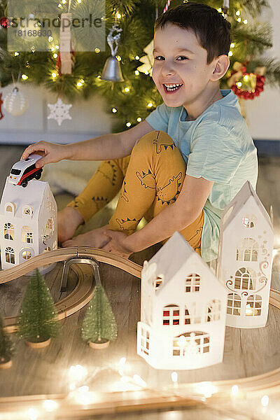 Fröhlicher Junge genießt mit Spielzeugeisenbahn und Hausmodell die Weihnachtsdekoration zu Hause