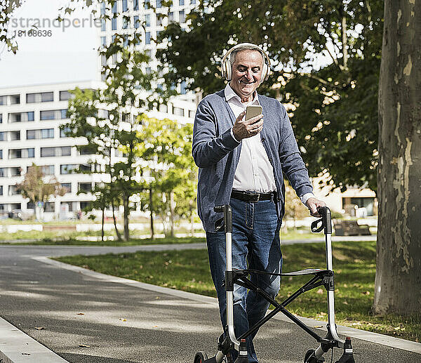Lächelnder älterer Mann  der sein Mobiltelefon benutzt und über drahtlose Kopfhörer Musik hört  während er mit einem Rollator auf dem Fußweg läuft