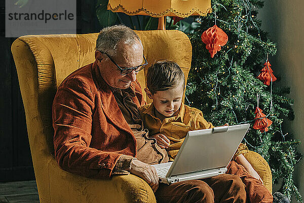 Großvater und Junge sitzen im Sessel und benutzen Laptop am Weihnachtsbaum