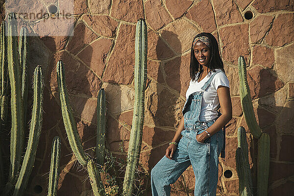 Lächelnde junge Frau  die an einem sonnigen Tag vor einer Wand mit einem Kaktus steht