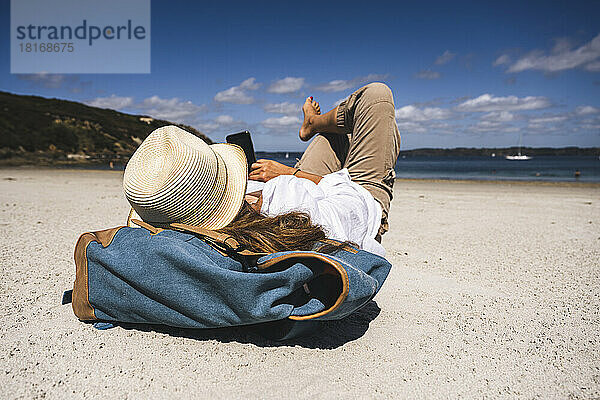 Reife Frau mit Hut und Smartphone am Strand liegend