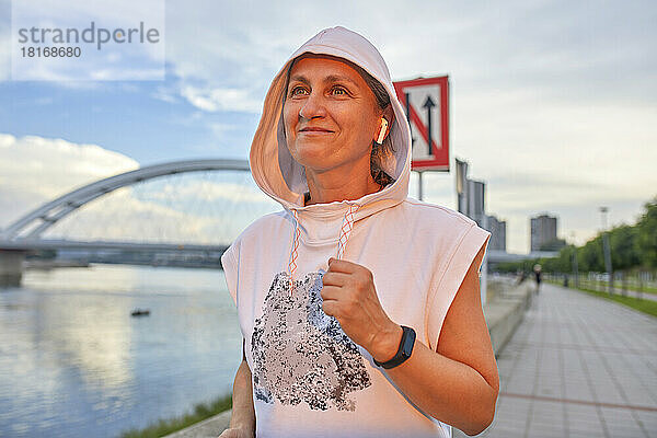 Lächelnde reife Frau mit kabellosen In-Ear-Kopfhörern joggt an der Promenade