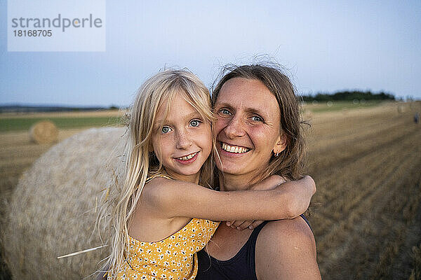 Lächelndes Mädchen mit blonden Haaren umarmt Mutter auf dem Feld