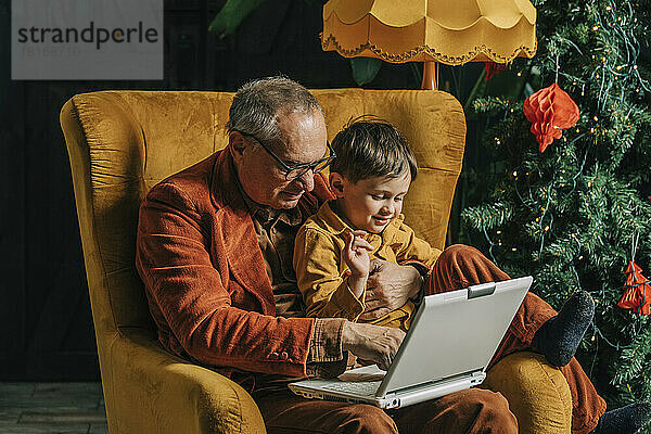 Großvater und Enkel sitzen im Sessel und benutzen Laptop  im Hintergrund der Weihnachtsbaum