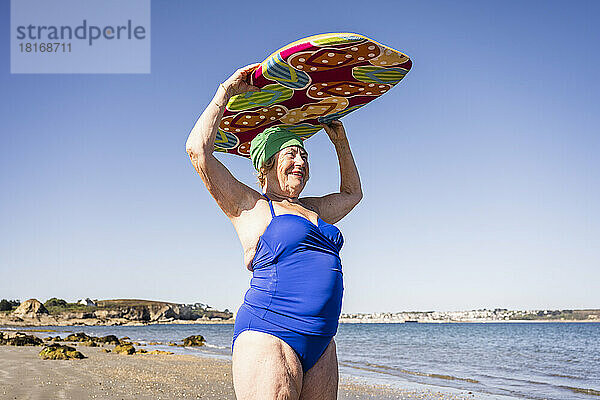 Glückliche ältere Frau trägt Surfbrett über dem Kopf am Strand