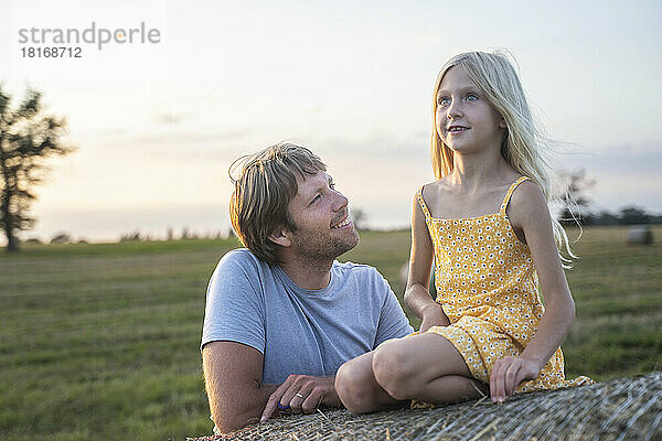 Lächelnder Mann schaut Tochter an  die auf Heuhaufen sitzt