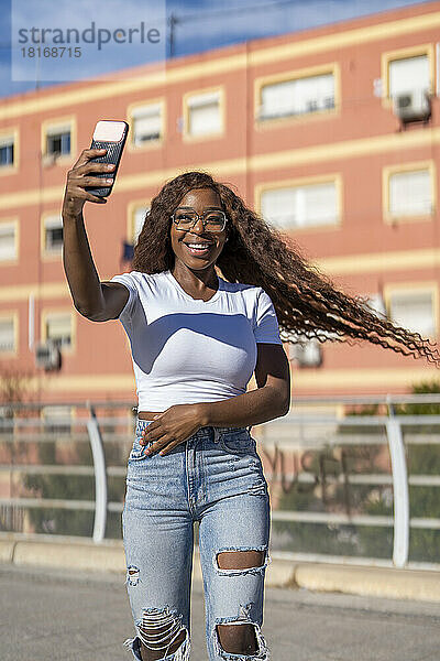 Glückliche junge Frau mit zerzausten Haaren  die auf dem Fußweg ein Selfie mit dem Smartphone macht