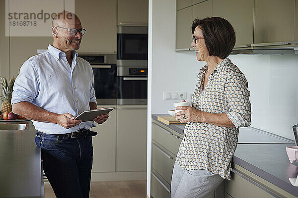 Glücklicher älterer Mann hält Tablet-PC in der Hand und steht mit Frau in der heimischen Küche