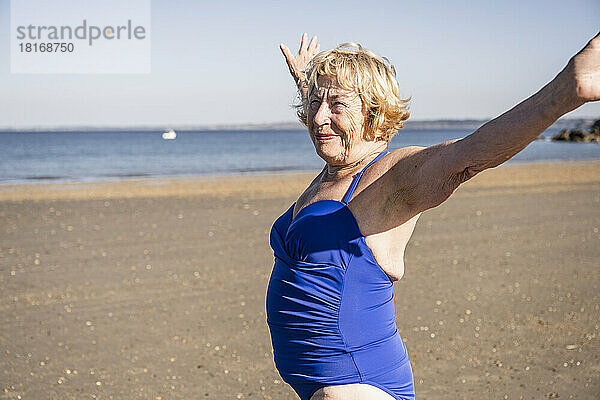 Lächelnde ältere Frau mit ausgestreckten Armen steht am Strand