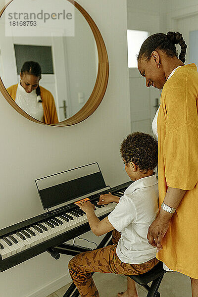 Lächelnde Frau schaut ihrem Sohn zu  der zu Hause Klavier spielt