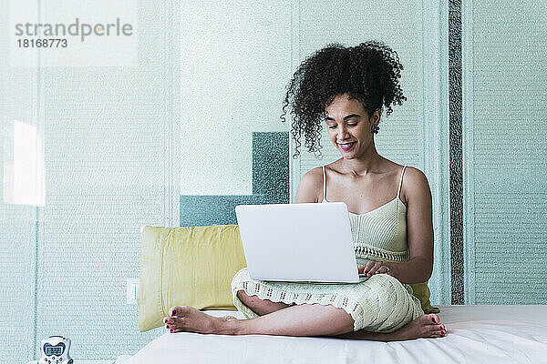 Lächelnde Frau mit lockigem Haar  die zu Hause im Bett einen Laptop benutzt