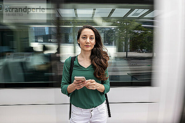 Lächelnde Frau steht mit Smartphone an der Straßenbahnhaltestelle