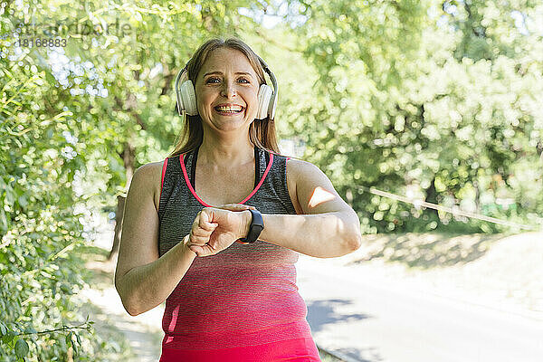 Lächelnde Frau mit kabellosen Kopfhörern  die im Park den Puls auf dem Fitness-Tracker überprüft