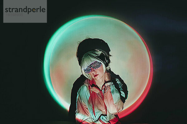Woman in neon eyeglasses standing against black background with spot lit