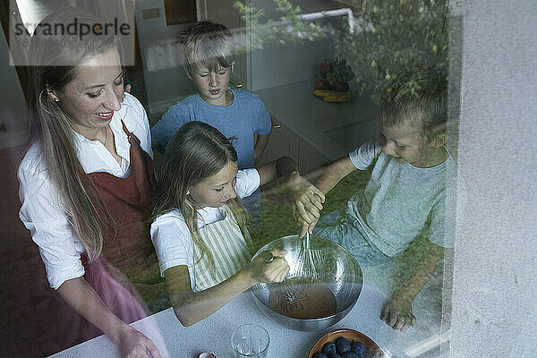 Mutter und Kinder mischen Teig für Kuchen in der Küche  gesehen durch Fensterglas