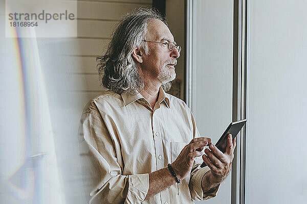 Nachdenklicher älterer Mann mit Tablet-PC am Fenster zu Hause