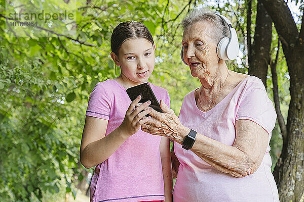 Enkelin hilft Urgroßmutter im Park mit Smartphone und Kopfhörern
