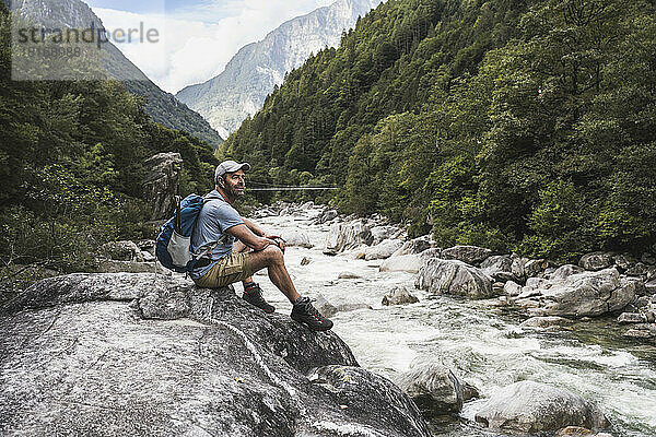 Nachdenklicher reifer Mann mit Mütze sitzt auf einem Felsen am Fluss