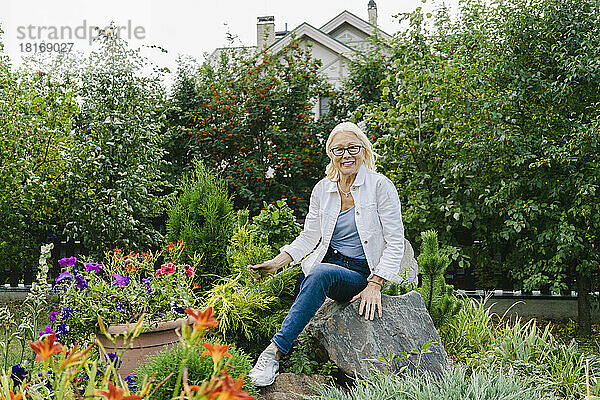 Lächelnde ältere Frau sitzt auf einem Felsen inmitten von Pflanzen im Garten