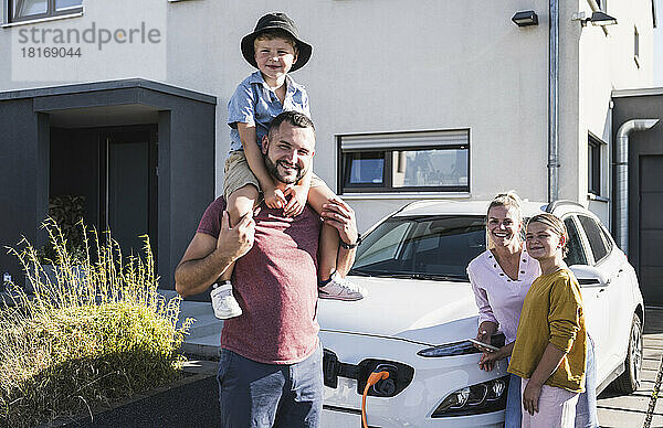 Glückliche Familie steht vor dem Einfamilienhaus und lädt Elektroauto auf