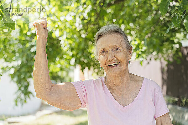 Glückliche ältere Frau  die vor einem Baum im Park ihre Muskeln spielen lässt