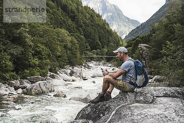 Reifer Mann sitzt mit Smartphone auf einem Felsen am Fluss
