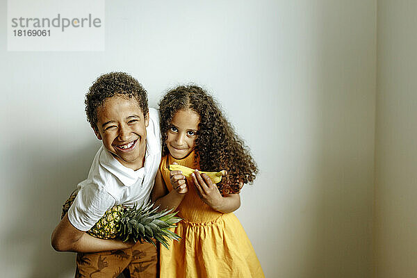 Glücklicher Bruder mit Ananas umarmt Schwester und hält Banane vor der Wand