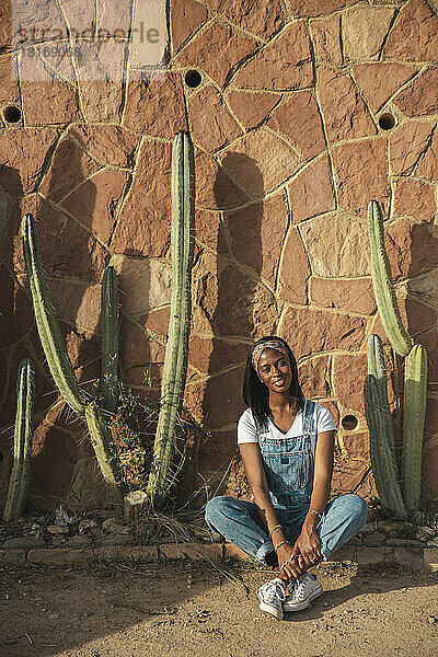 Lächelnde junge Frau sitzt an einem sonnigen Tag vor der Wand
