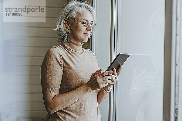 Reife Frau mit grauen Haaren nutzt Tablet-PC am Fenster zu Hause
