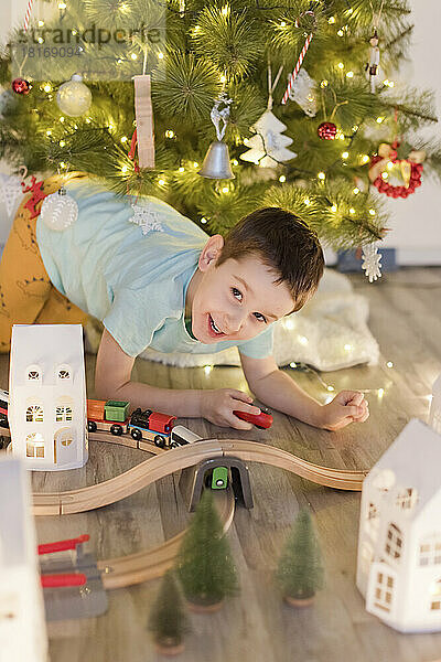 Fröhlicher Junge  der zu Hause mit einer Miniatur-Spielzeugeisenbahn am Weihnachtsbaum spielt