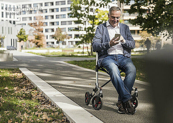 Lächelnder älterer Mann sitzt auf einem Rollator und nutzt sein Smartphone
