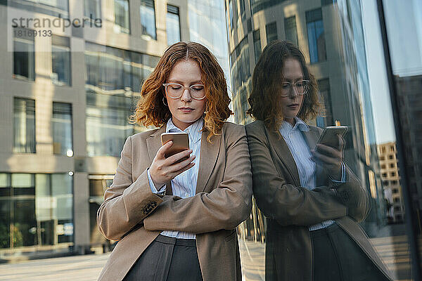 Geschäftsfrau mit Brille benutzt Smartphone und lehnt an Glaswand