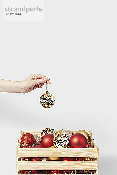 Woman's hand holding golden bauble over box with colorful balls against white background